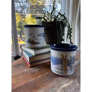 May include: Two white ceramic mugs with blue rims and handles. One mug features a cabin in a snowy landscape, the other has skis and a snowy mountain scene. The mugs are on a wooden table.