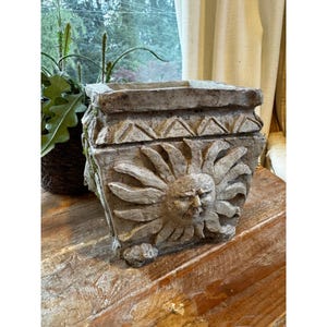 May include: A square, weathered concrete planter with a sun face design. The planter has a textured surface, decorative trim, and small feet. The neutral color and detailed carving give it a rustic, antique appearance. Perfect for adding a unique touch to any garden or patio.