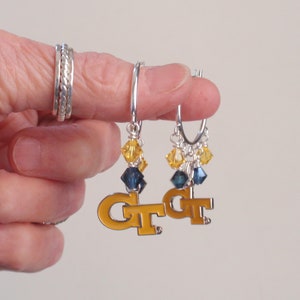 May include: Silver hoop earrings with yellow and blue crystal accents. Each earring features a gold-colored "GT" charm. The earrings are held up by a person's finger, with silver rings on the finger.
