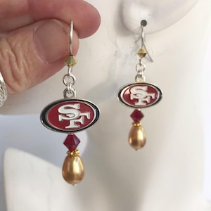 May include: A pair of silver earrings with a red, white, and gold San Francisco 49ers logo charm, a red crystal, and a gold pearl.