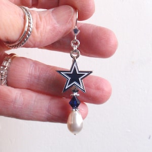 May include: Silver dangle earrings with a blue and white star charm and a white pearl drop. The star charm has a silver outline and a blue center with a white star in the center.