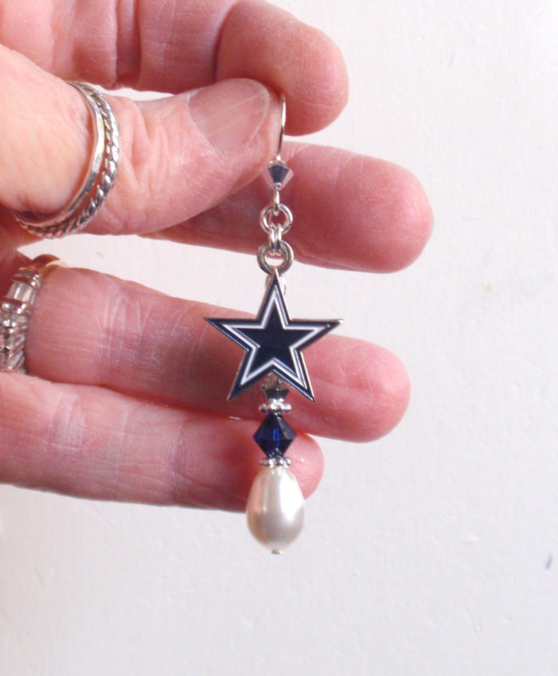 May include: Silver dangle earrings with a blue and white star charm and a white pearl drop. The star charm has a silver outline and a blue center with a white star in the center.