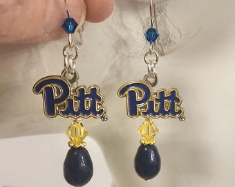 May include: A pair of dangle earrings featuring the word "Pitt" in blue and gold. Each earring has a blue teardrop-shaped bead and a yellow crystal accent. The earrings are suspended from silver-colored hooks.