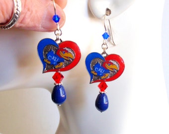 May include: Heart-shaped earrings with a red and blue design, featuring a bird logo and the letters "KU". The earrings have a silver hook, a blue crystal, a red crystal, and a blue teardrop bead.