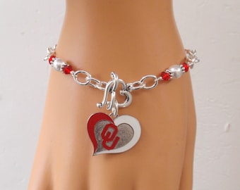 May include: Silver-toned bracelet with a toggle clasp. The bracelet features red and silver beads, and a heart-shaped charm with a red and white design. The charm has the letter "O" inside.