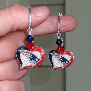 May include: A pair of heart-shaped earrings with a silver finish. Each earring features a red and navy blue heart design with a silver logo. The earrings are accented with red and blue beads and have a hoop closure.
