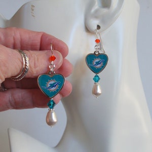 May include: A pair of silver heart-shaped earrings with a teal blue background and a white dolphin logo. The earrings have a single clear crystal and a white pearl hanging from each heart.