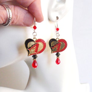 May include: A pair of dangle earrings featuring a red, black, and gold heart-shaped charm with a number one on it. The charm is attached to a silver chain with a red crystal bead at the bottom.
