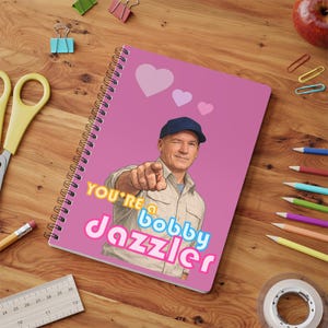 May include: Pink spiral notebook with the text "YOU'RE a Bobby Jazzler" and an image of a man pointing. The notebook has a pink cover with three hearts and a silver spiral binding. Pencils, scissors, and paper clips are on the wooden surface.