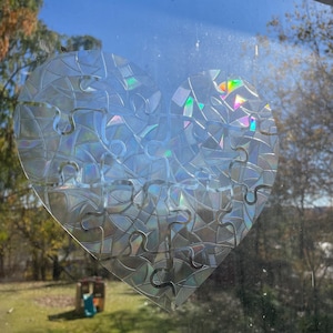 Op de afbeelding: Een hartvormige raamsticker gemaakt van iriserende puzzelstukjes. De sticker is geplaatst op een raam met uitzicht op een groen gazon en bomen.