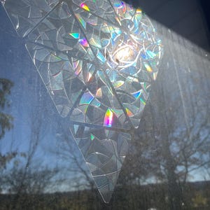 Puede incluir: Una pegatina de ventana triangular iridiscente con un patrón geométrico. La pegatina está hecha de un material transparente con un efecto arcoíris.