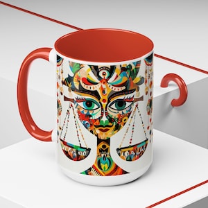 May include: A white ceramic mug with a red rim and handle. The mug features a colorful illustration of a stylized face with scales on either side. The face is made up of geometric shapes and patterns in shades of red, orange, yellow, green, blue, and purple.