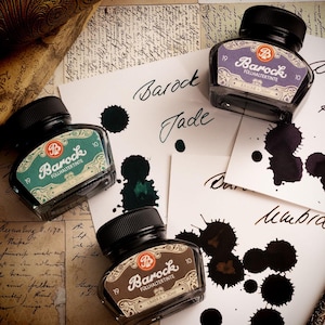 May include: An overhead shot features four bottles of Barock fountain pen ink in various colors, with ink splotches on paper. The bottles have black caps and labels with ornate designs. The paper has handwritten text and the ink colors are Jade, Umbra, and Kaviar.