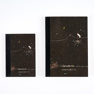 Pode incluir: Dois cadernos retangulares com lombadas pretas e capas castanho-escuras com uma ilustração de céu noturno com lua crescente, estrelas, um gato num fio e o horizonte de uma cidade. O texto "ほぼ日の方眼ノート" está impresso na parte inferior das capas.
