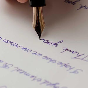 May include: A close-up of a fountain pen with a gold nib writing on a white piece of paper. The pen is being held by a hand, and the paper has blue ink on it. The text on the paper reads "Thank you" and "I hope that we can return to normal soon."