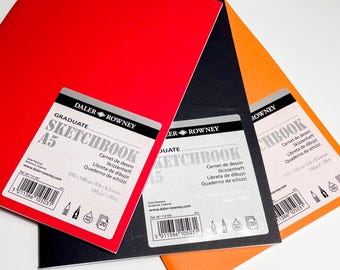 May include: Three A5 sketchbooks in red, black, and orange, with white labels. The labels read "GRADUATE SKETCHBOOK A5" and include measurements of 210 x 148 mm / 5.9 x 8.3 inches. The brand name "DALER ROWNEY" is also visible.
