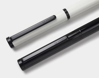 May include: Two pens, one white and one black, are displayed at an angle. Each pen has a black clip with the text "le chat" printed on it. The white pen has a black band near the top. The pens are set against a light gray background.