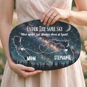 May include: An oval plaque featuring a night sky background with a galaxy, stars, and a forest silhouette. It displays the text "Under The Same Sky" and "Miles apart, but always close at heart," with outlines of California and Florida connected by a heart-shaped dotted line, labeled "MOM" and "STEPHANIE."
