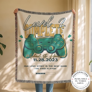 May include: Cream-colored woven blanket with a teal and gold video game controller design. Text reads "Level 2 Complete" with a couple's names and date. The blanket also says "Our love story is the best game I've ever played!"