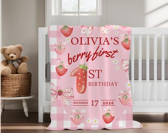Puede incluir: Una manta rosa suave con un borde de cuadros blancos y un patrón de fresas con lazos rosas. El texto dice OLIVIA'S berry first 1ST BIRTHDAY NOVEMBER 17 2026, con una fresa grande con forma de número uno en el centro.