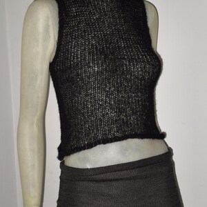 Op de afbeelding: Een zwarte, mouwloze gebreide top met een hoge halslijn en een bijpassende zwarte rok. De top heeft een licht gerimpelde rand aan de onderkant. De rok is een minirok. De outfit wordt op een mannequin getoond.