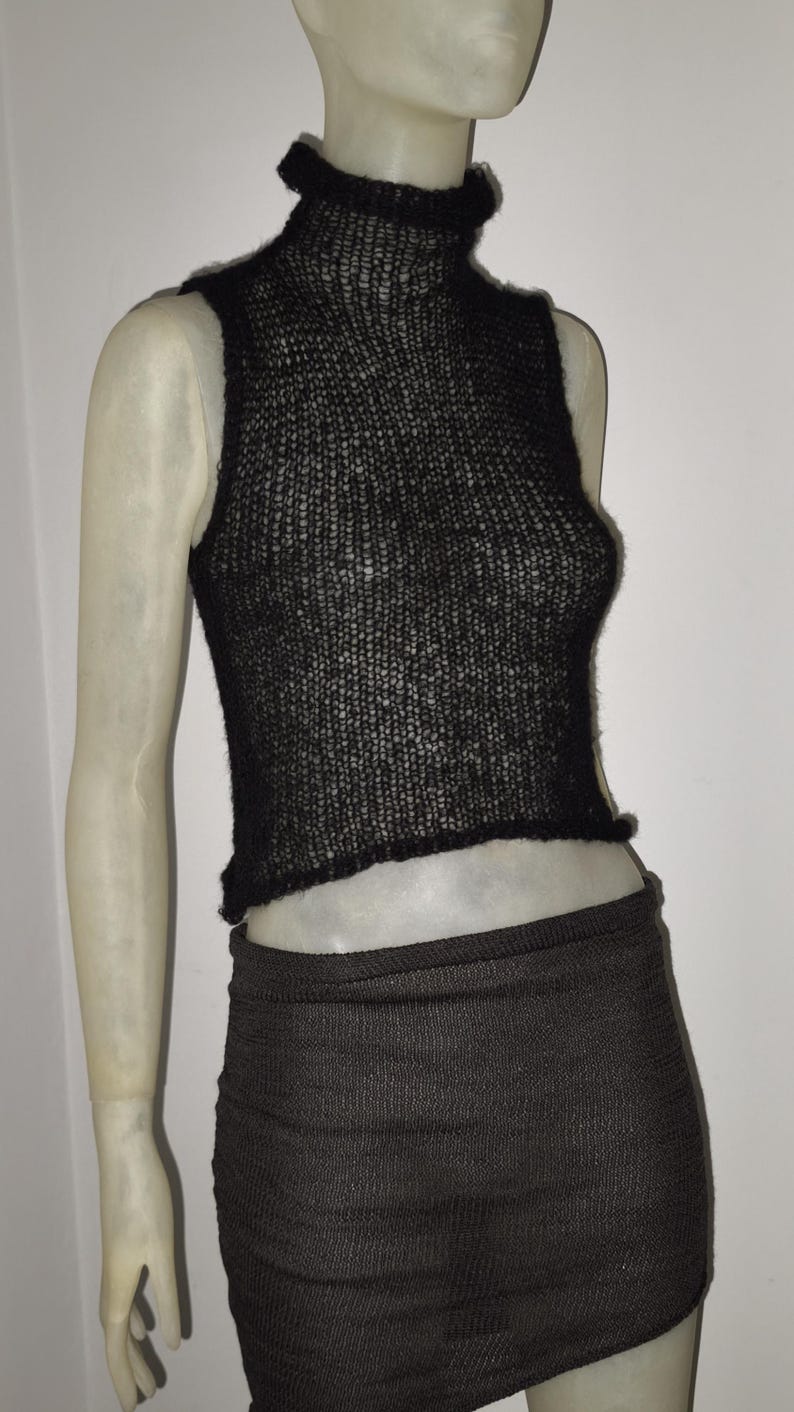 Op de afbeelding: Een zwarte, mouwloze gebreide top met een hoge halslijn en een bijpassende zwarte rok. De top heeft een licht gerimpelde rand aan de onderkant. De rok is een minirok. De outfit wordt op een mannequin getoond.