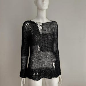 May include: A black long-sleeve jumper featuring a loose, open-knit construction with intentional distressed holes throughout the fabric. The garment includes a lace-up detail at the upper chest area, creating a cutout effect in the knit pattern.