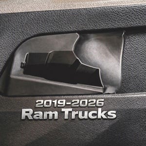 May include: A black textured plastic interior storage compartment designed for a vehicle door panel. The surface features the embossed silver text 2019-2026 Ram Trucks. The component has a molded recessed pocket with a smooth interior finish.