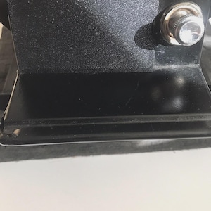 May include: Close-up of a black metal object with a textured surface and a shiny, silver knob. The object has a rectangular shape with a glossy finish on the lower portion. The background is a white surface.