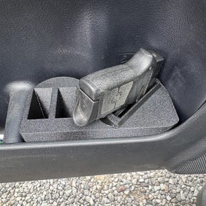 May include: Black handgun resting in a custom-fit, black plastic holder inside a car door panel. The holder is designed to securely cradle the firearm, with additional compartments visible. The car door panel is black.