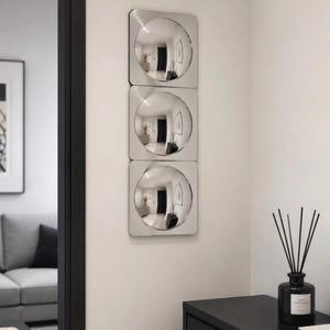 Peut inclure: Une unité murale verticale composée de trois panneaux carrés argentés, chacun intégrant un miroir convexe circulaire. Les surfaces réfléchissantes présentent une vue déformée de la pièce environnante, créant un effet visuel répétitif.