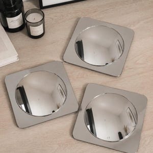 May include: Three square silver-toned coasters, each featuring a circular, reflective, convex center. The coasters have rounded corners and a smooth, metallic finish, creating a polished appearance that reflects the surrounding environment.