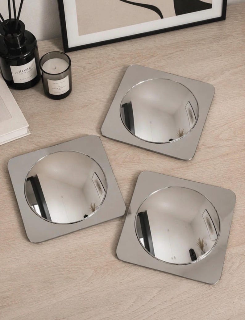 May include: Three square silver-toned coasters, each featuring a circular, reflective, convex center. The coasters have rounded corners and a smooth, metallic finish, creating a polished appearance that reflects the surrounding environment.