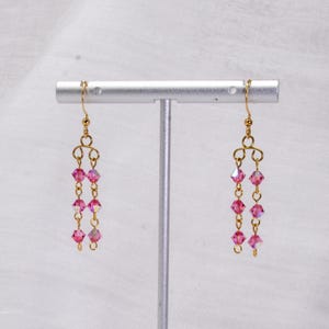 May include: A pair of gold dangle earrings with pink crystal beads. The earrings are hanging from a silver stand.