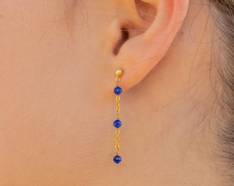 Puede incluir: Pendientes colgantes dorados con tres cuentas azul oscuro suspendidas de una delicada cadena. La imagen en primer plano resalta el contraste entre el oro y el azul de las joyas.