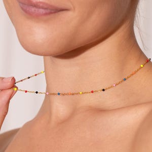 Könnte beinhalten: Eine zarte goldene Halskette mit kleinen, farbigen Perlen. Die Kette hat eine feine Kette und ist mit einer Reihe von leuchtenden, mehrfarbigen Perlen verziert, darunter Rot, Gelb, Blau und Schwarz. Die Halskette wird gehalten, um ihr Design zu zeigen.