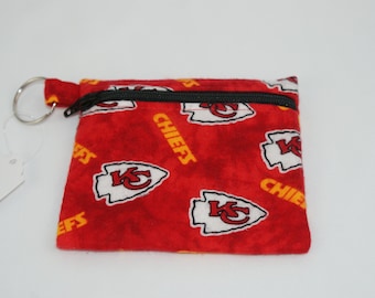 May include: A small, red fabric coin purse with a black zipper and a silver key ring. The purse is patterned with white and yellow logos and the word "CHIEFS".