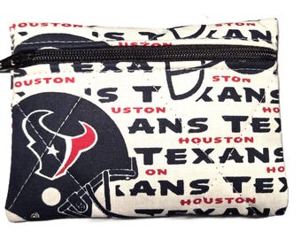May include: A small, rectangular zippered pouch featuring a repeating pattern of the words "HOUSTON TEXANS" and a navy blue and red Houston Texans helmet graphic on a cream background. The zipper is black.