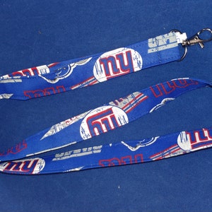 May include: Blue lanyard with a white and red New York Giants logo repeated on the fabric. The lanyard has a silver metal clip.