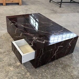 May include: A rectangular coffee table with a dark brown marble-like finish and white veining. The table has a drawer that is pulled out, revealing a white interior. The table is in a room with a concrete floor.
