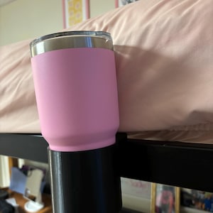May include: A pink and black insulated tumbler holder attached to a black metal bed frame. The holder is designed to keep a tumbler upright and secure.