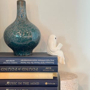 Puede incluir: Un jarr&oacute;n de cer&aacute;mica moteado turquesa y una peque&ntilde;a figura de fantasma blanco leyendo un libro se encuentran sobre una pila de libros. Los libros son de varios tonos de azul y dorado. Los t&iacute;tulos de los libros incluyen "Sundays Will Never Be the Same".