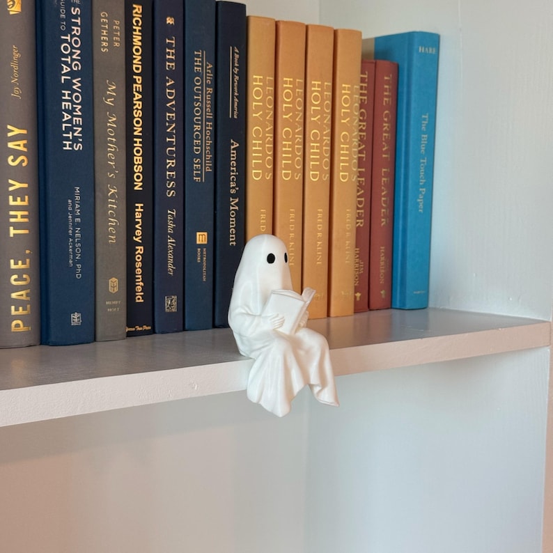 Puede incluir: Una figura de fantasma de cer&aacute;mica blanca leyendo un libro, posada en una estanter&iacute;a blanca. El fantasma tiene ojos negros y est&aacute; envuelto en una s&aacute;bana fluida. Una colecci&oacute;n de libros de tapa dura de varios colores forma el fondo.