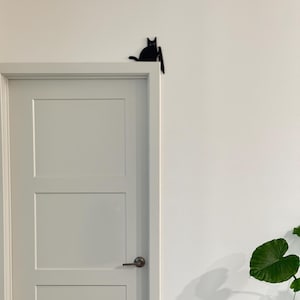 May include: A black silhouette of a cat perched on the top corner of a white door frame. The cat figure is flat and solid black, positioned as if sitting on the edge of the door casing, with its tail extending slightly along the top horizontal edge.
