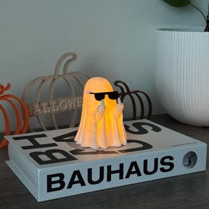 Puede incluir: L&aacute;mpara con forma de fantasma brillante, con gafas de sol y mostrando el dedo coraz&oacute;n. Se encuentra sobre un libro con la palabra "BAUHAUS". Decoraciones de calabazas de madera y una maceta blanca en el fondo.