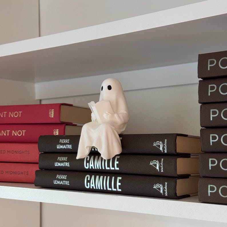 Puede incluir: Una figura de fantasma blanco de cer&aacute;mica leyendo un libro, posada sobre una pila de libros marrones oscuros. Los libros tienen el t&iacute;tulo "CAMILLE" en letras blancas. La estanter&iacute;a tambi&eacute;n muestra otros libros con cubiertas rojas.