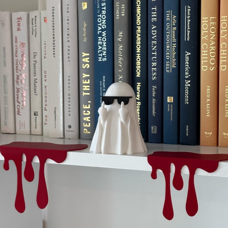 Puede incluir: Una figura de fantasma blanco con gafas de sol, mostrando el dedo medio, colocada en una estanter&iacute;a blanca. Decoraci&oacute;n de estanter&iacute;a roja goteando debajo. Libros con varios t&iacute;tulos en el fondo.