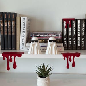 Puede incluir: Dos figuras de fantasmas blancos con gafas de sol est&aacute;n sobre una estanter&iacute;a blanca, flanqueadas por decoraciones de estanter&iacute;a rojas con forma de sangre. Se ven libros con t&iacute;tulos como "GENERATION NEXT OLI WHITE" y "NEGOTIAUCTIONS". Una peque&ntilde;a planta en maceta a&ntilde;ade un toque de verde.