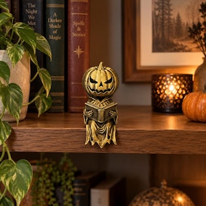 May include: A gold-colored figurine of a pumpkin-headed character with a jagged, carved face, wearing a hooded robe and holding an open book. The figure has a weathered, metallic finish and is designed to sit on the edge of a flat surface.