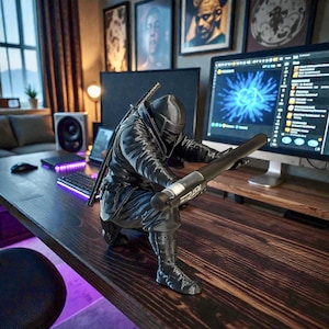 May include: A black ninja figurine kneeling on a wooden desk, holding a black cylindrical object with white text. The ninja is wearing a black mask and outfit, with a sword on its back. The desk has a computer and keyboard.